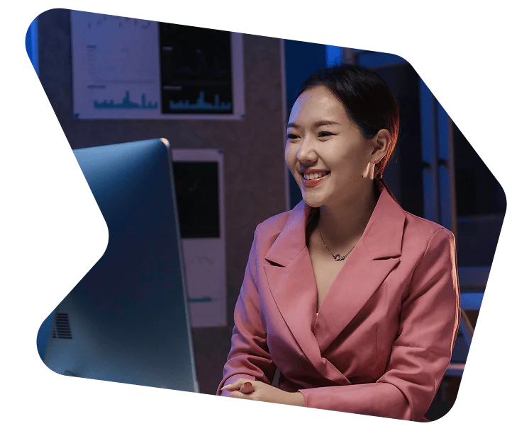 Woman in a pink blazer smiling and working at a computer in a dimly lit office